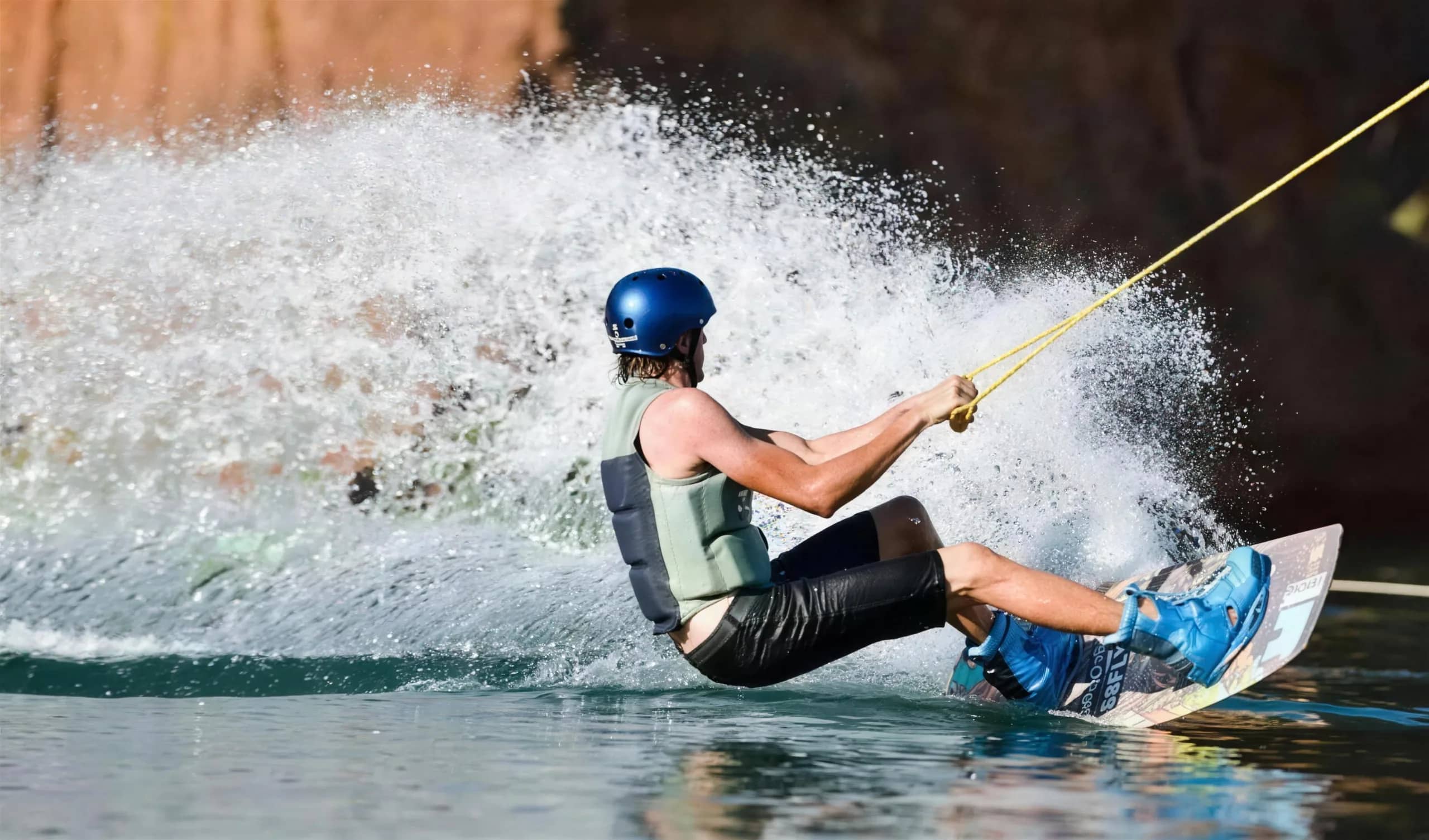 A wakeboarder in a helmet and vest carves across the water, throwing up a wall of spray.