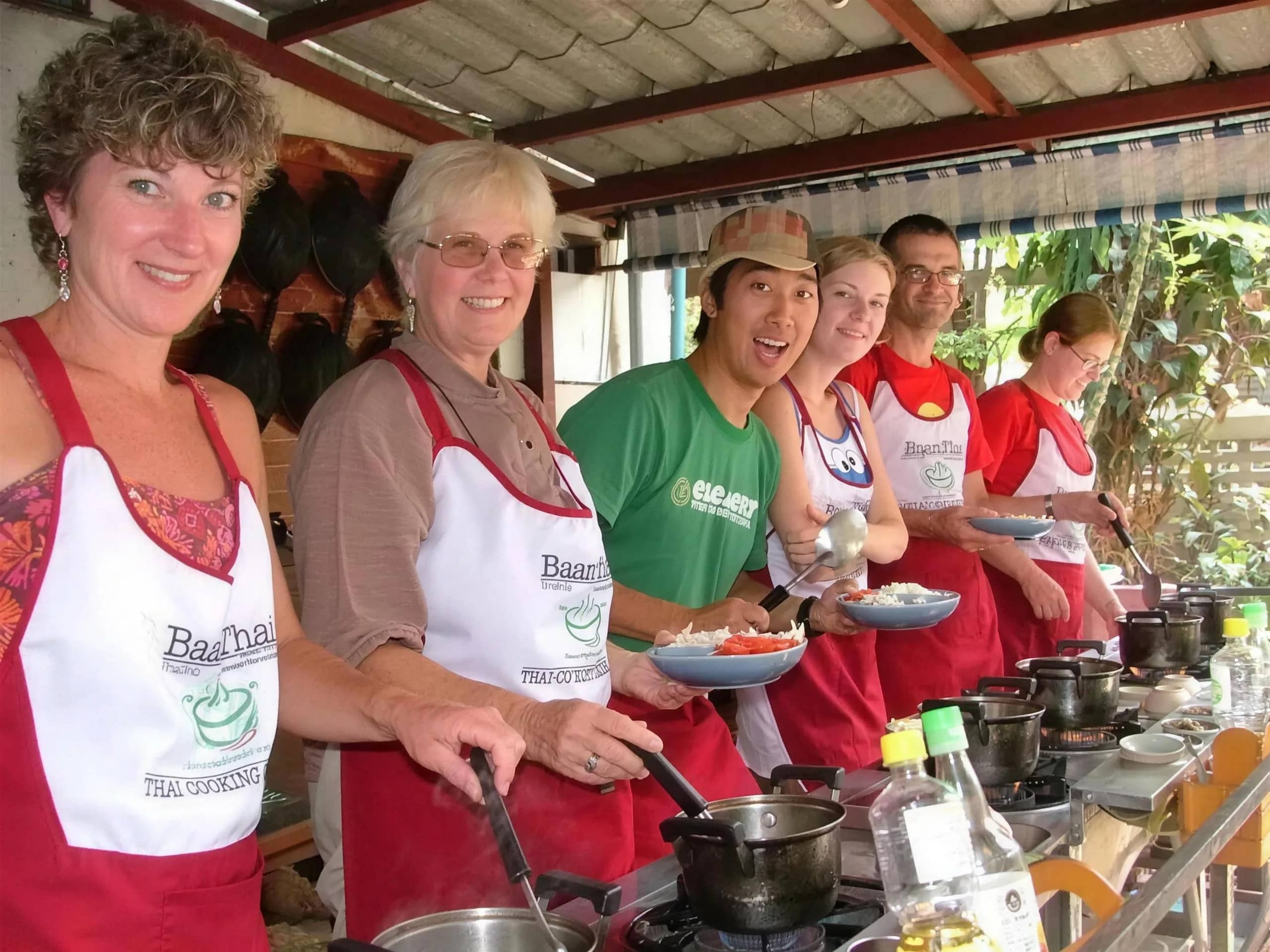 Guests in BaanThai aprons smile while cooking at a row of gas burners at the cooking school
