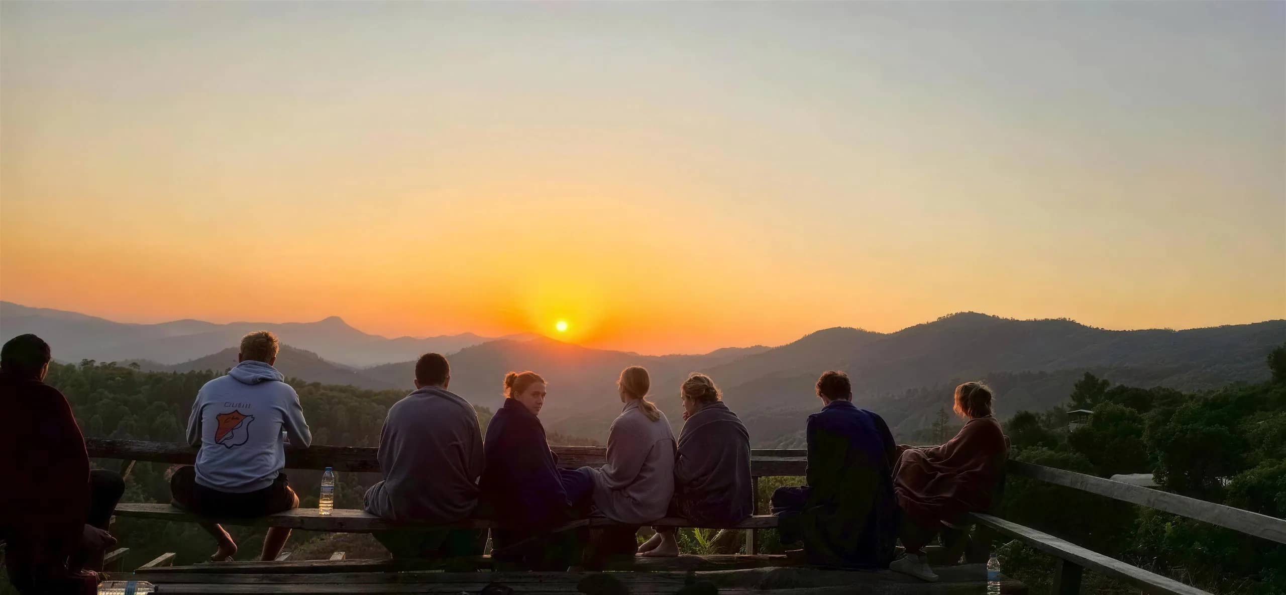 Trekkers sit on a viewpoint deck watching the sun set over layered mountains