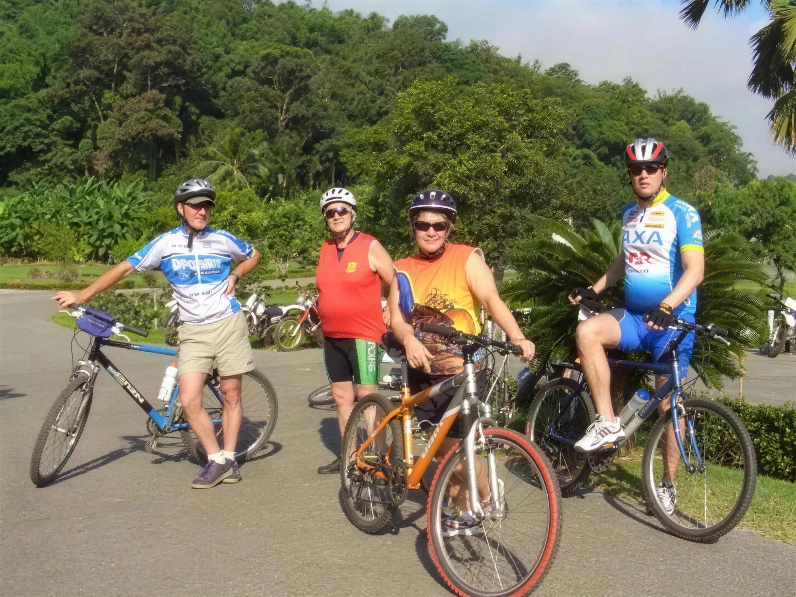 Four cyclists in helmets and jerseys pose with their bikes on a paved road beside green hills