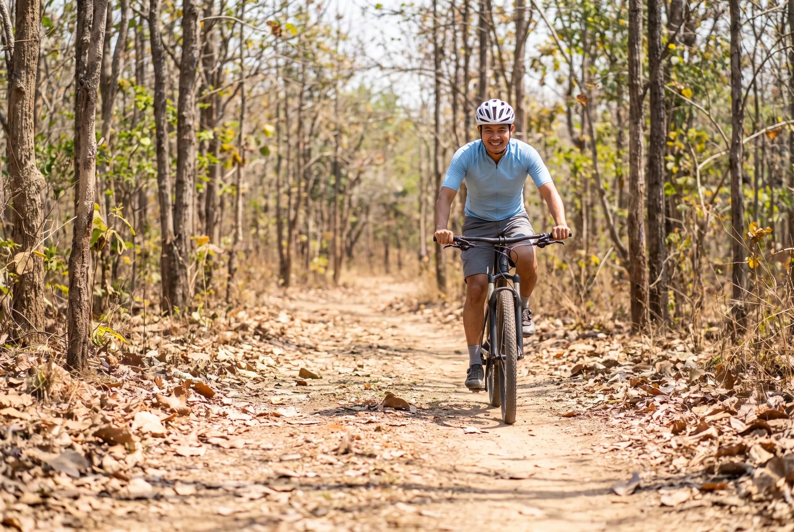 A cyclist in a light blue jersey rides towards the camera on a dirt trail lined with tall thin trees