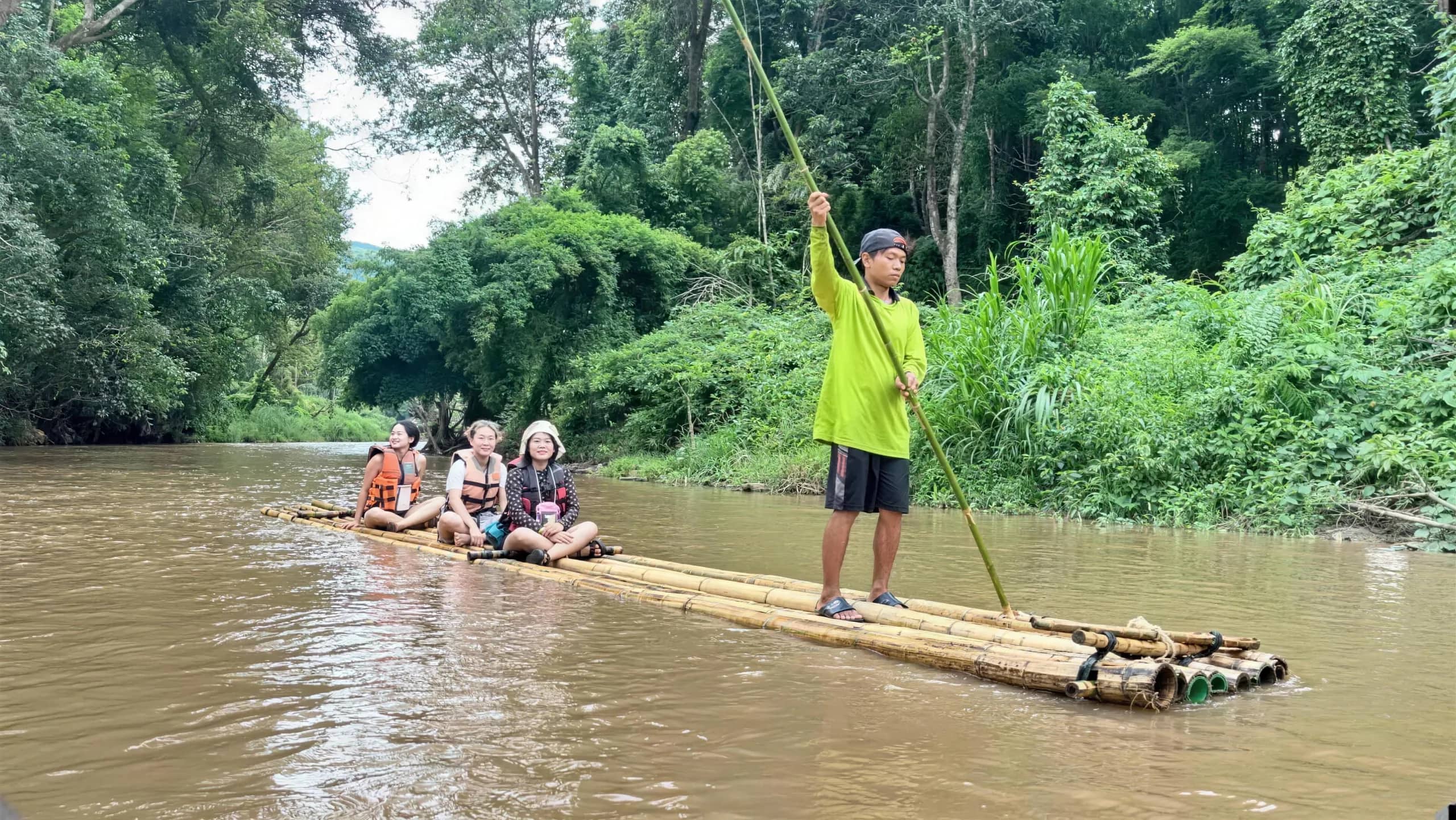 A guide in a green shirt stands at the back of a bamboo raft carrying visitors on a jungle river