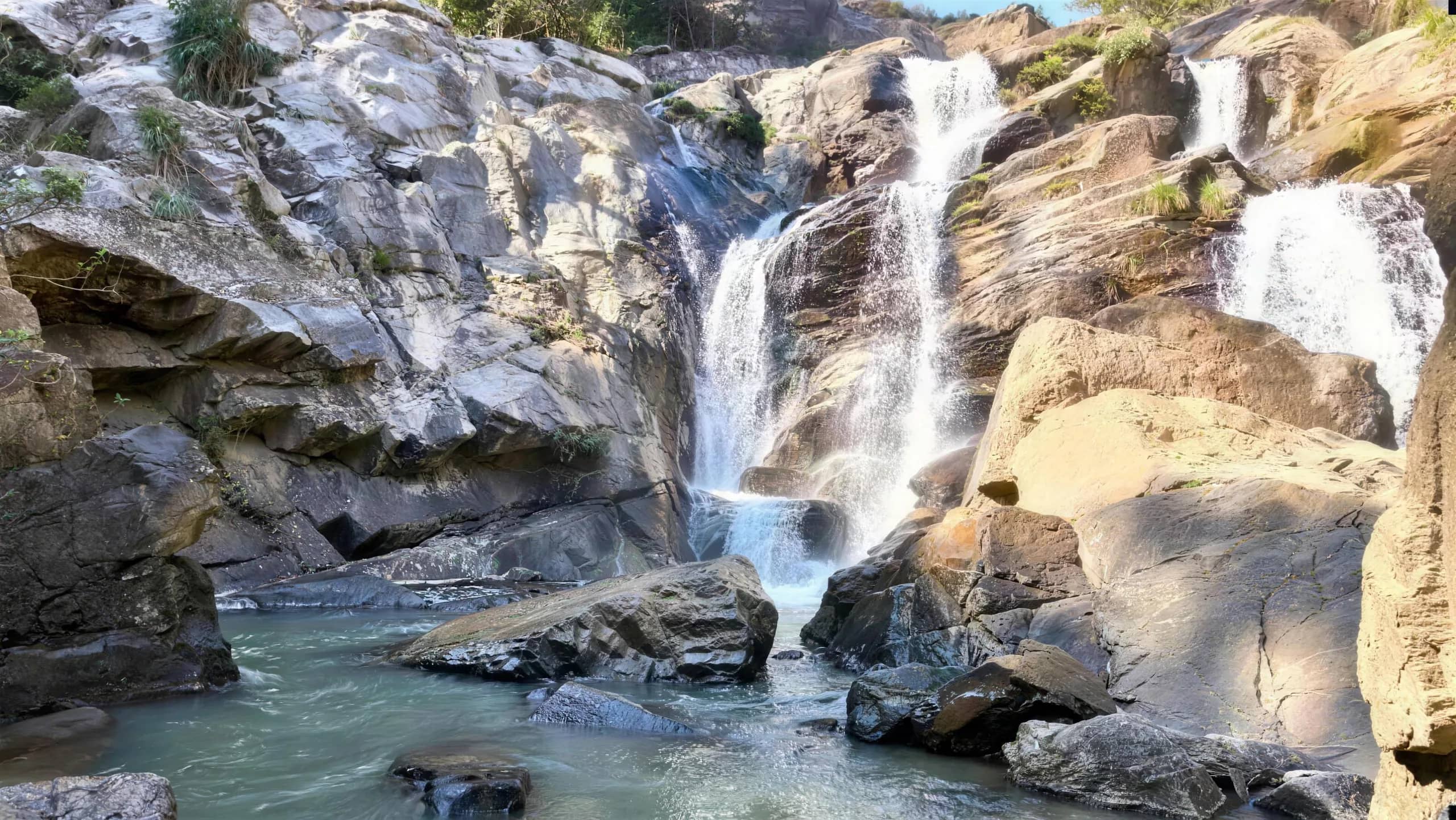 A waterfall pours down layered rock cliffs into a clear pool surrounded by boulders