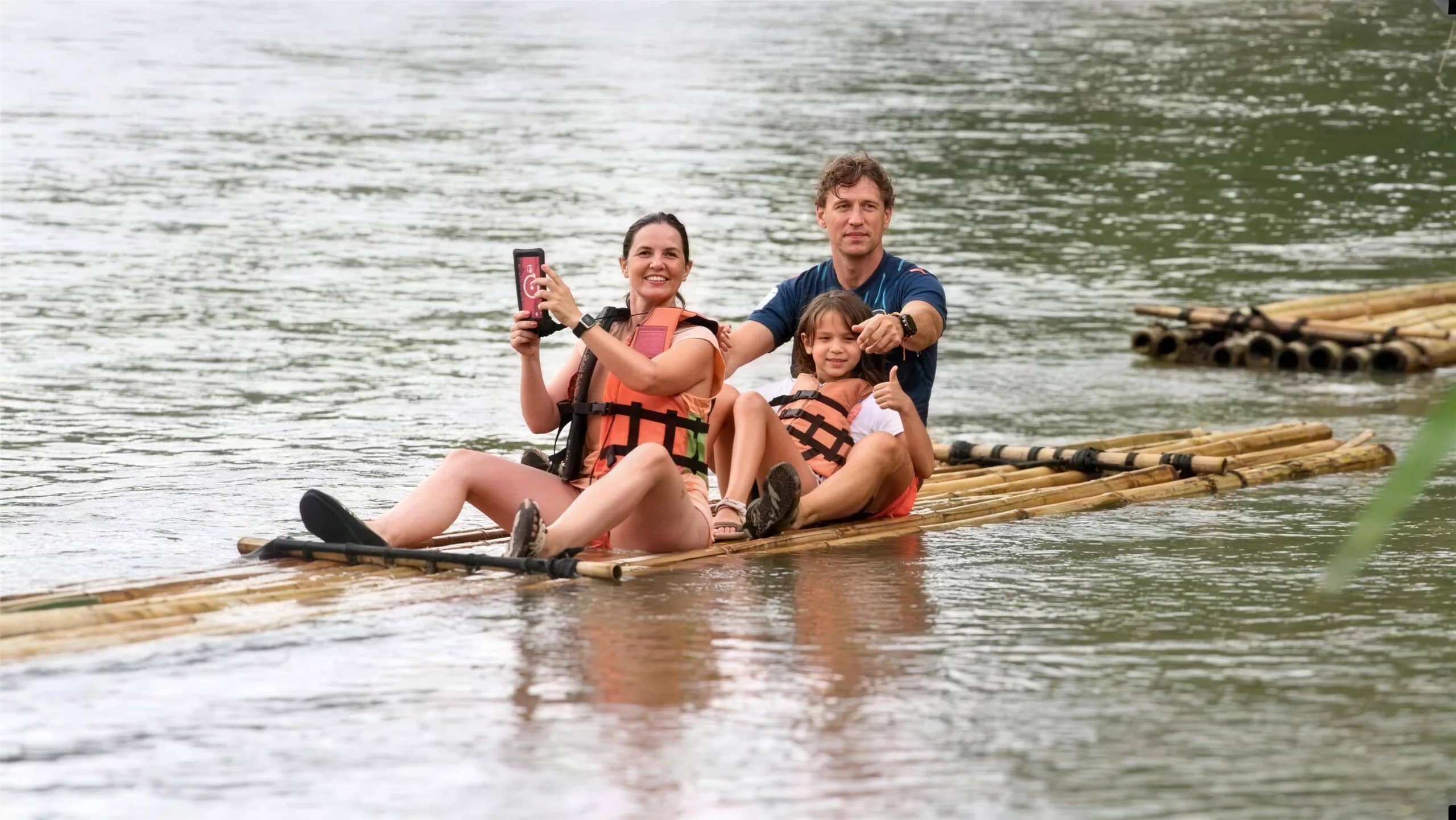 A smiling family in life jackets sits on a bamboo raft in the middle of a river