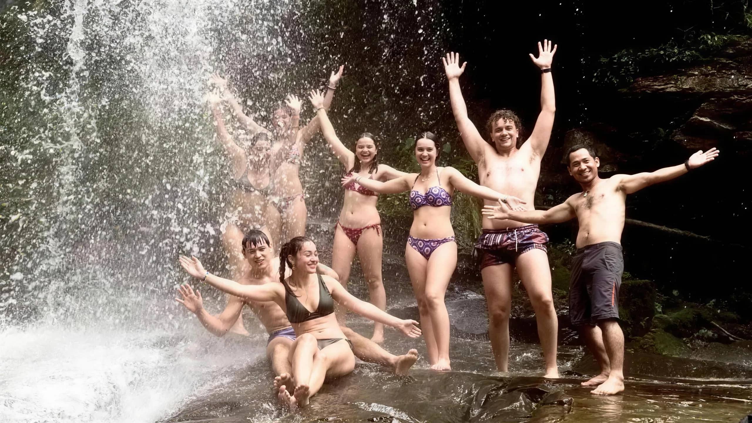 A group of visitors in swimwear raise their arms under the spray of a jungle waterfall