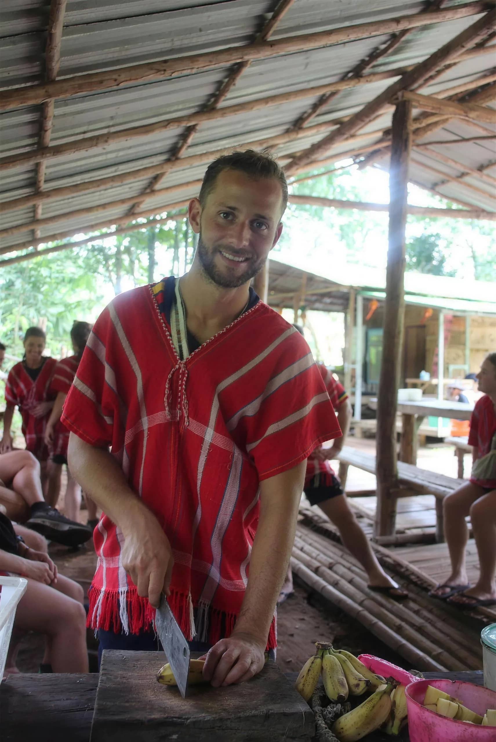 A visitor in a red Karen shirt prepares bananas at a wooden table under a shelter