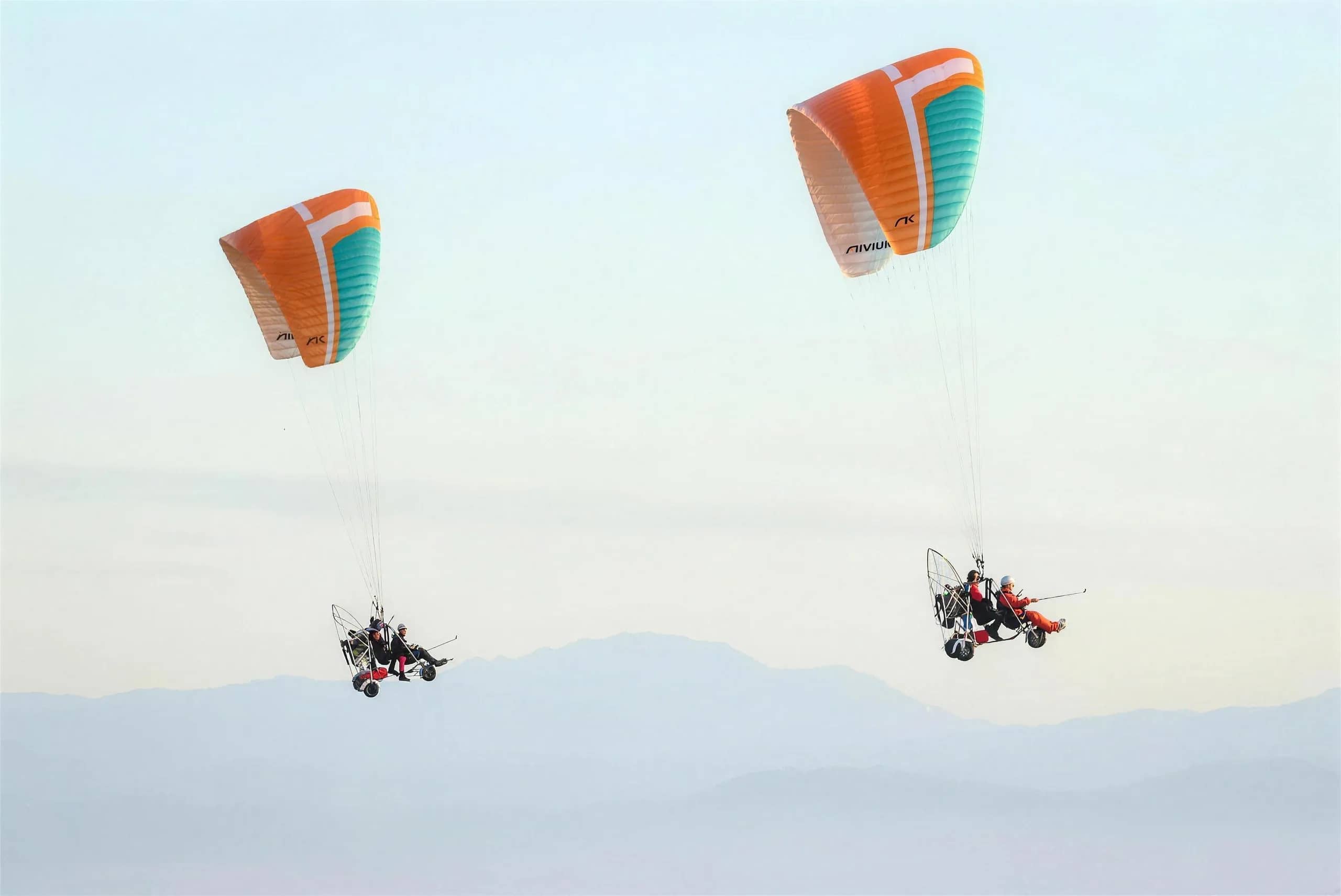 Two paramotors with orange and white wings fly against a pale sky above distant mountains