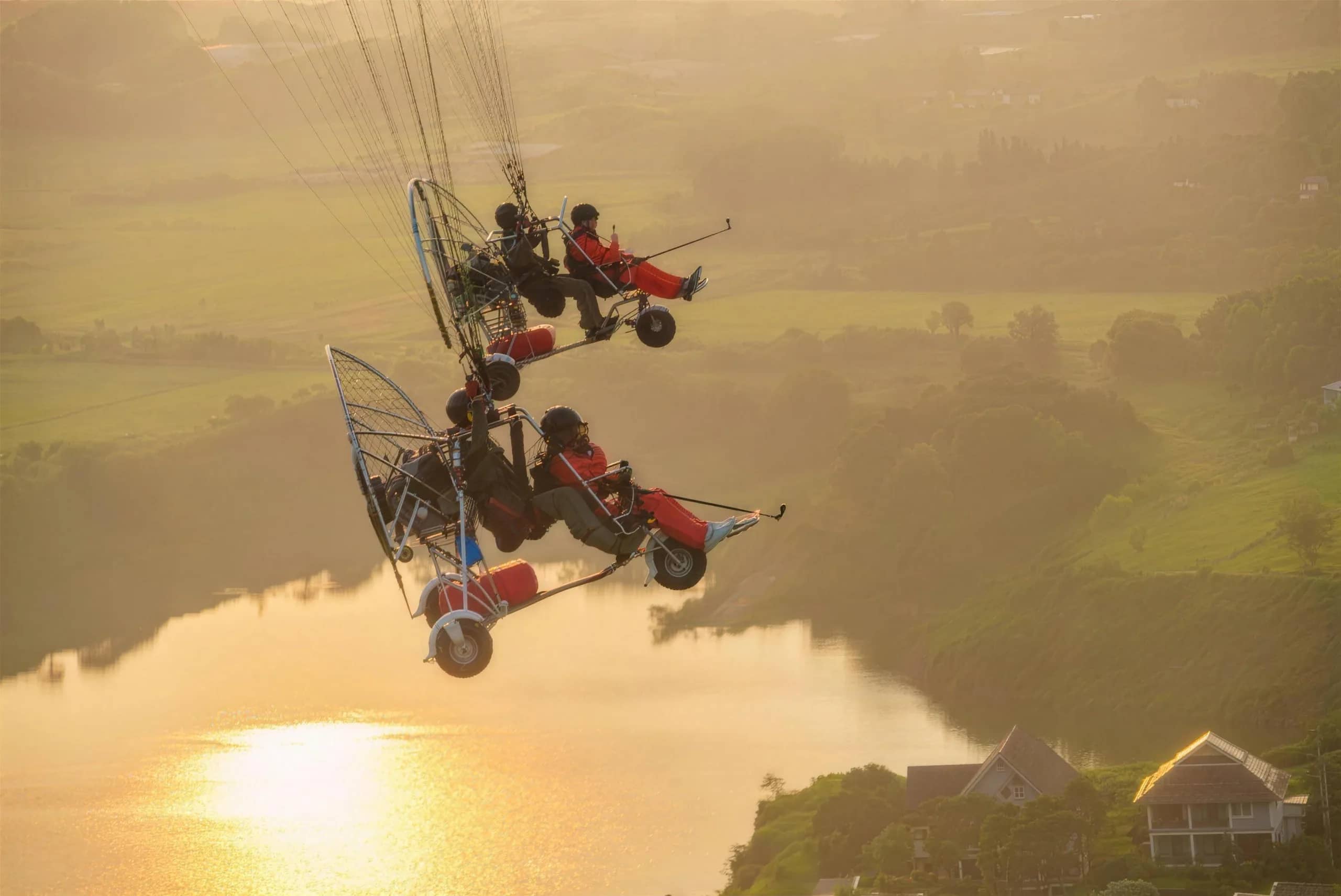 Two tandem paramotor trikes fly over a shining lake and green countryside at sunrise