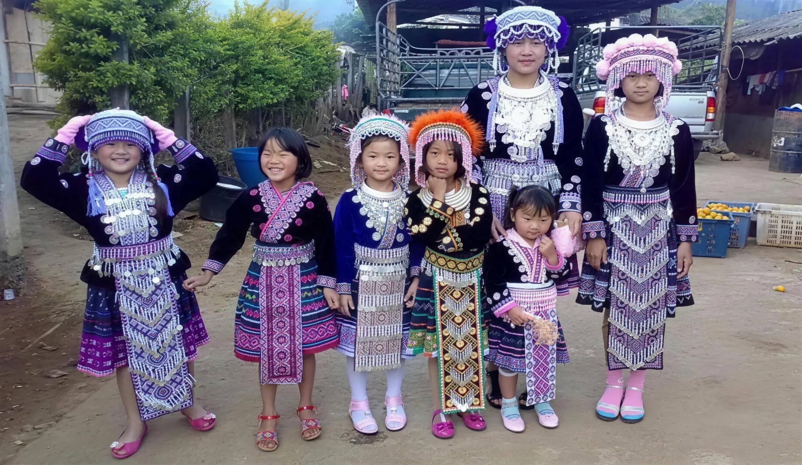 A group of children in embroidered Hmong costumes and headdresses poses in a village courtyard.