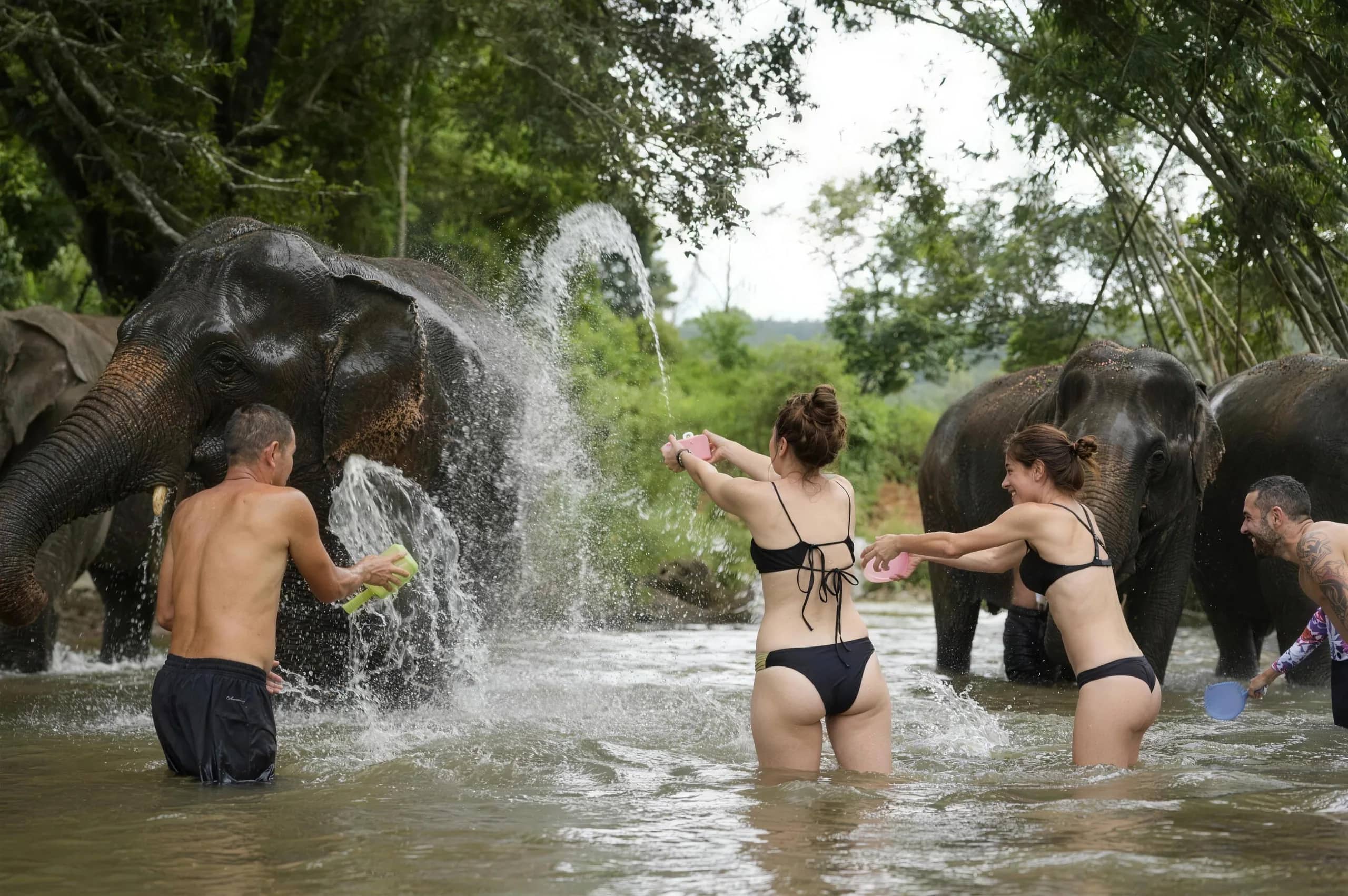Visitors throw water at elephants in a shallow river as the elephants spray water back