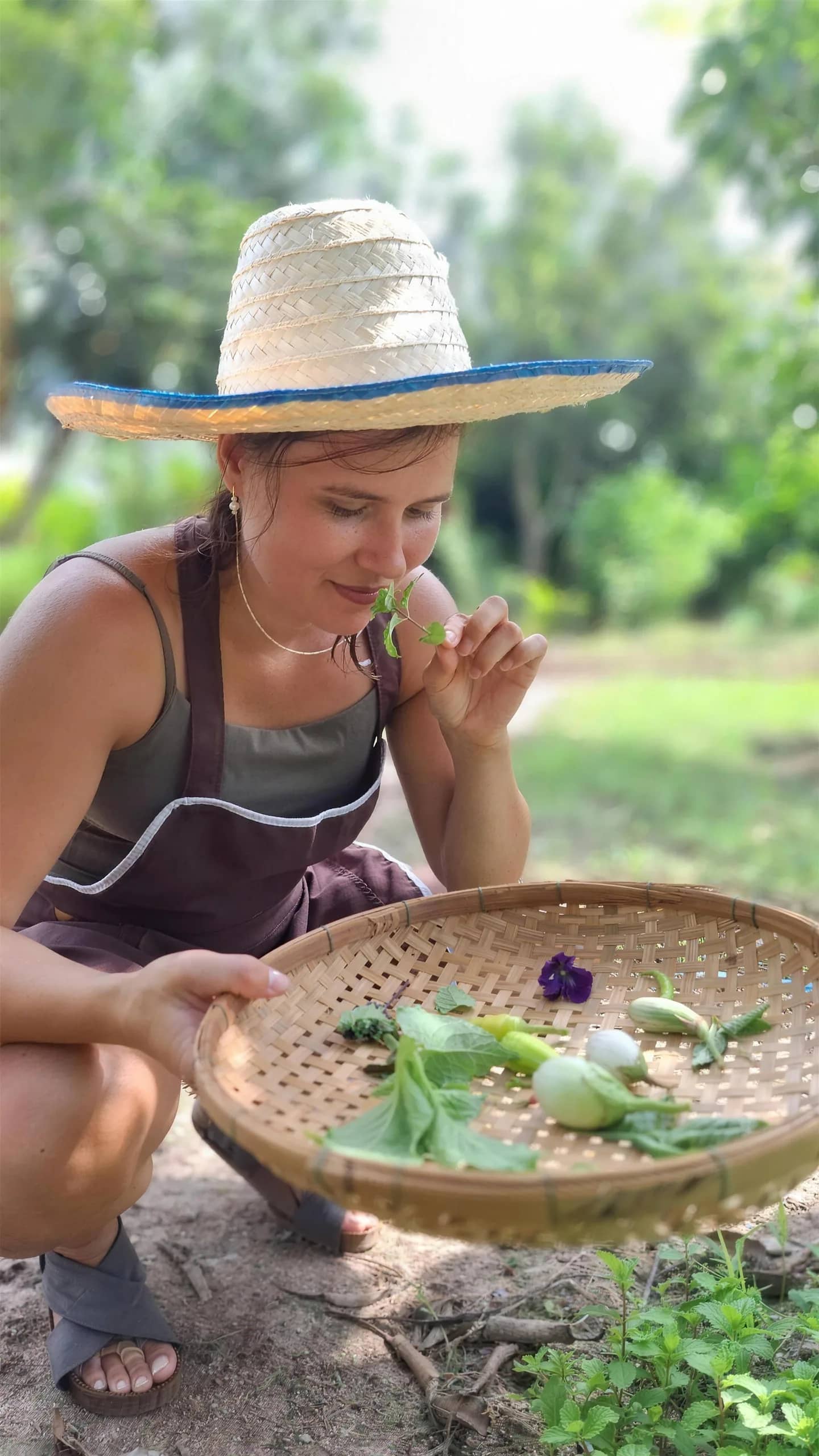 A woman in a straw hat smells fresh herbs in a woven basket while gathering ingredients.