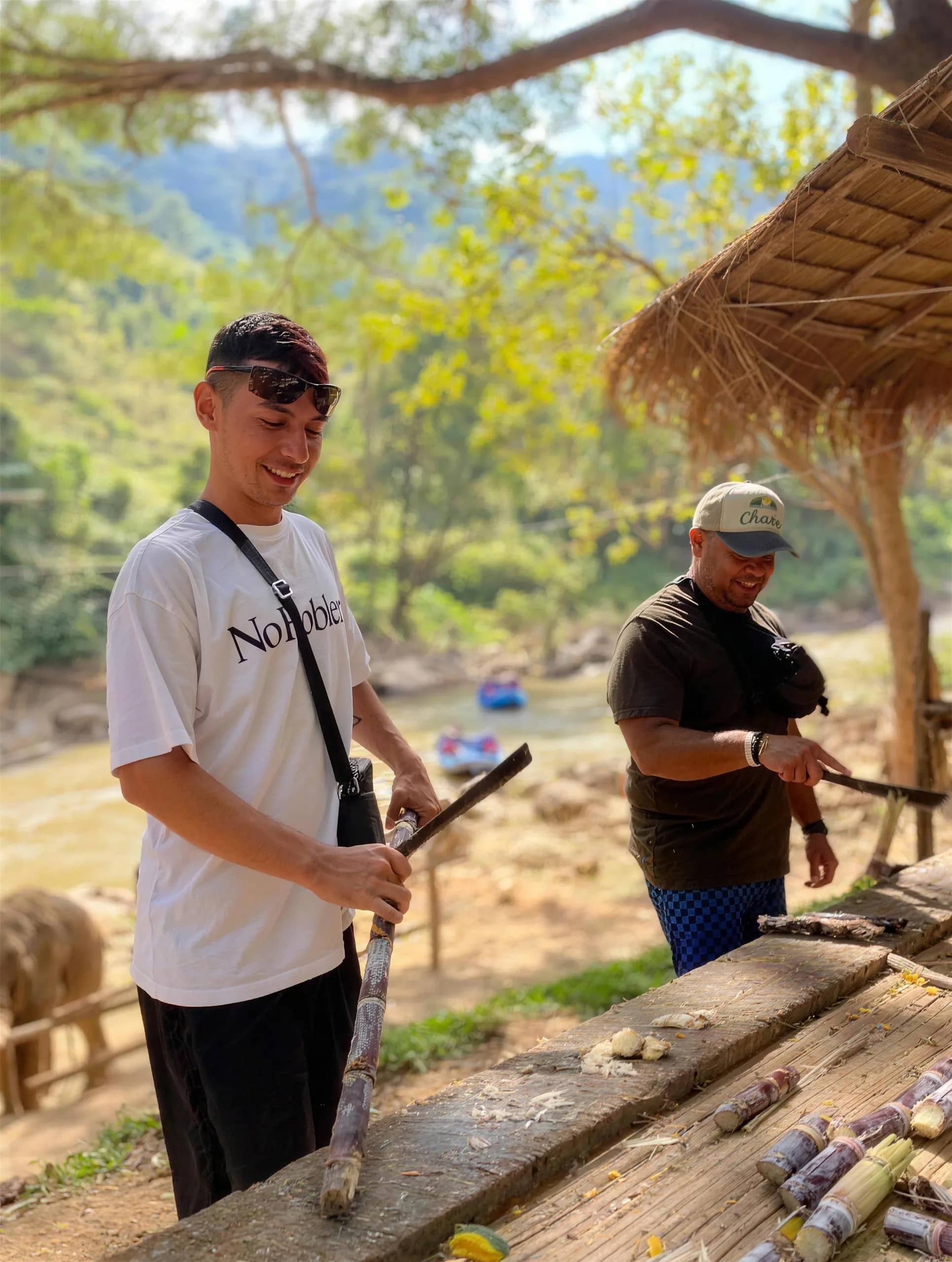 Two visitors chop sugarcane on a wooden table at a riverside elephant camp