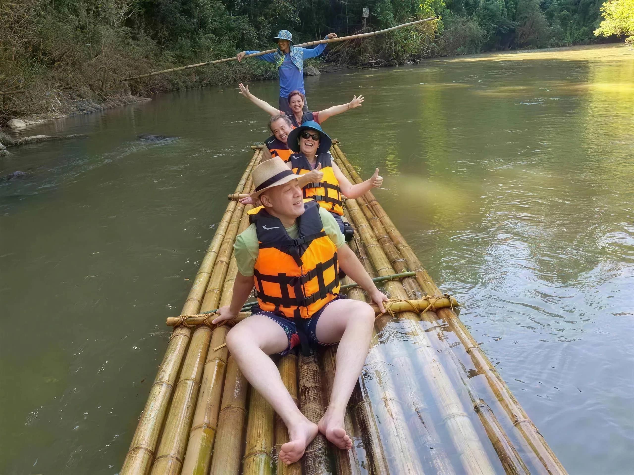 Guests in life jackets sit on a long bamboo raft as a guide poles it along a calm river.