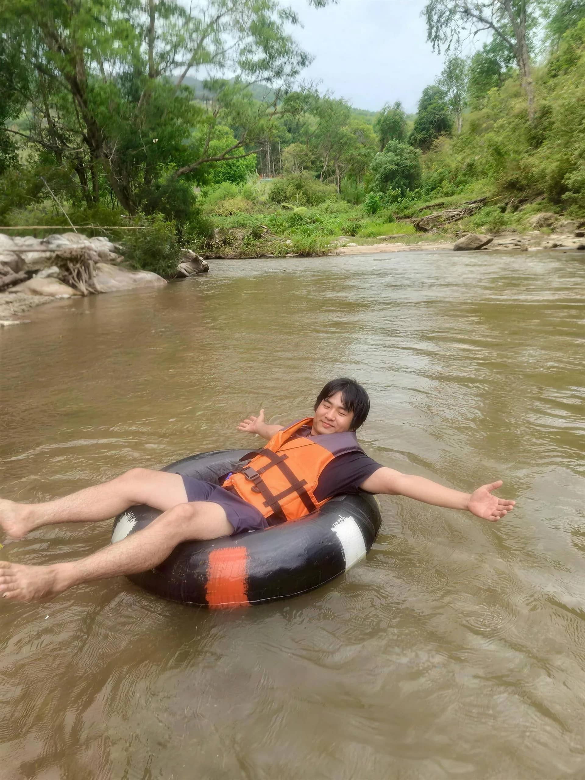 A smiling guest in a life jacket floats on a black tube down a brown jungle river