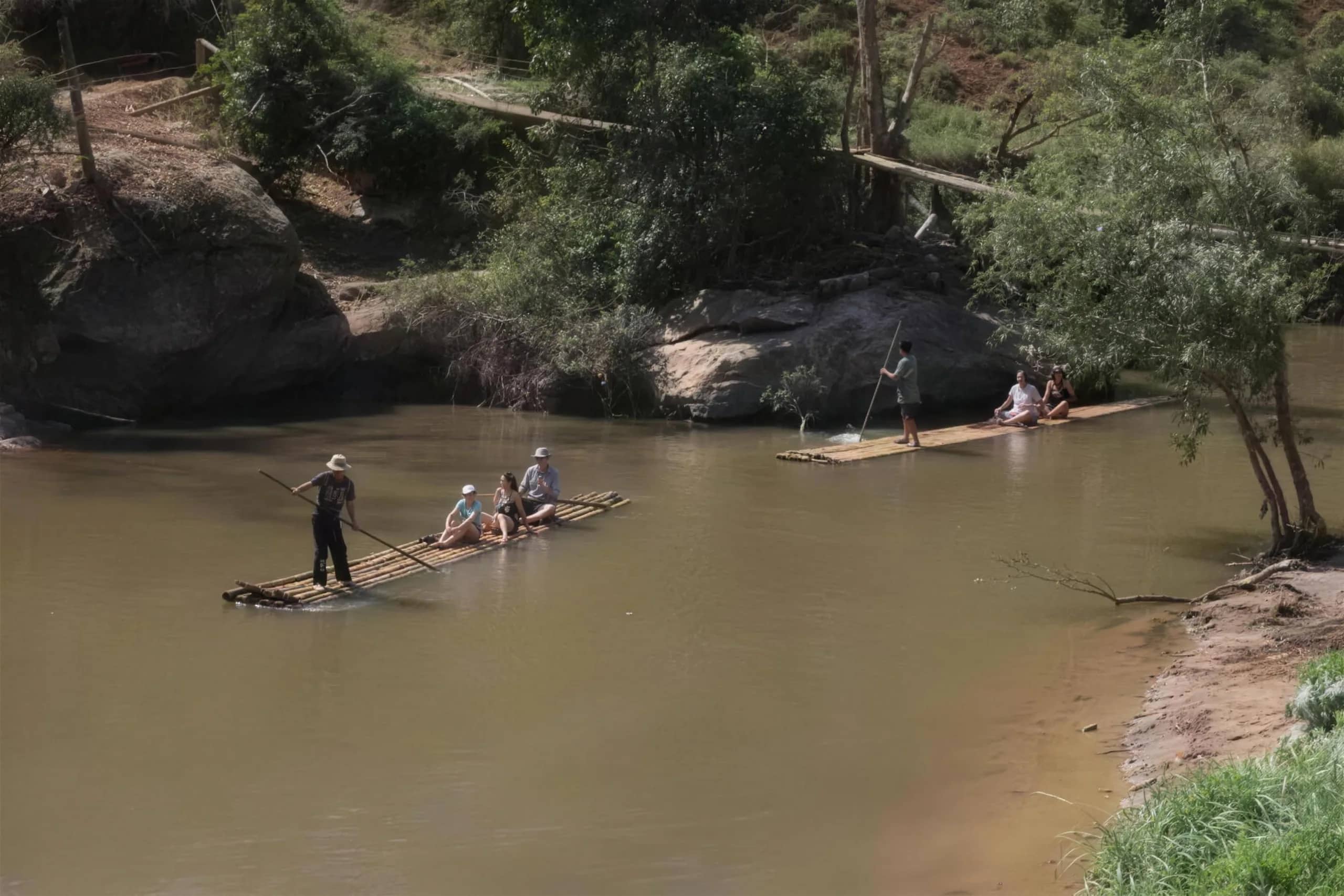 Two bamboo rafts with guides and visitors drift along a calm river below forested banks