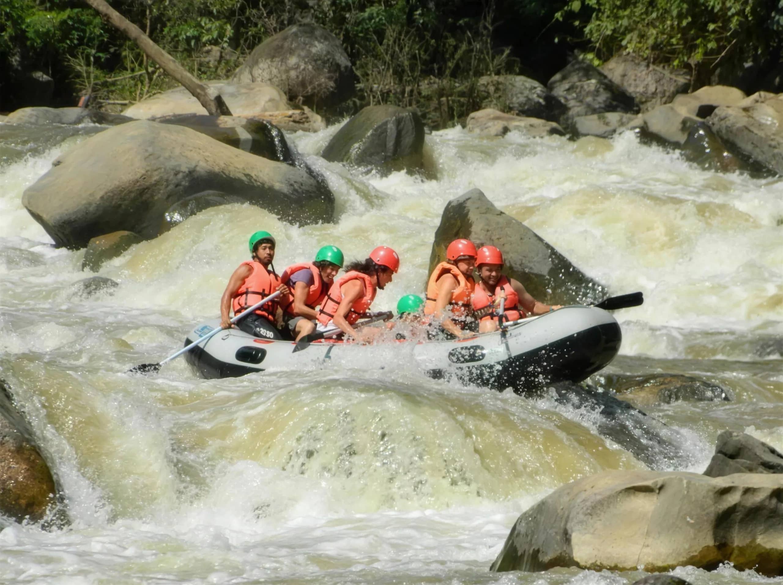 A raft crew in helmets and life jackets paddles through foaming rapids between boulders near Chiang Mai