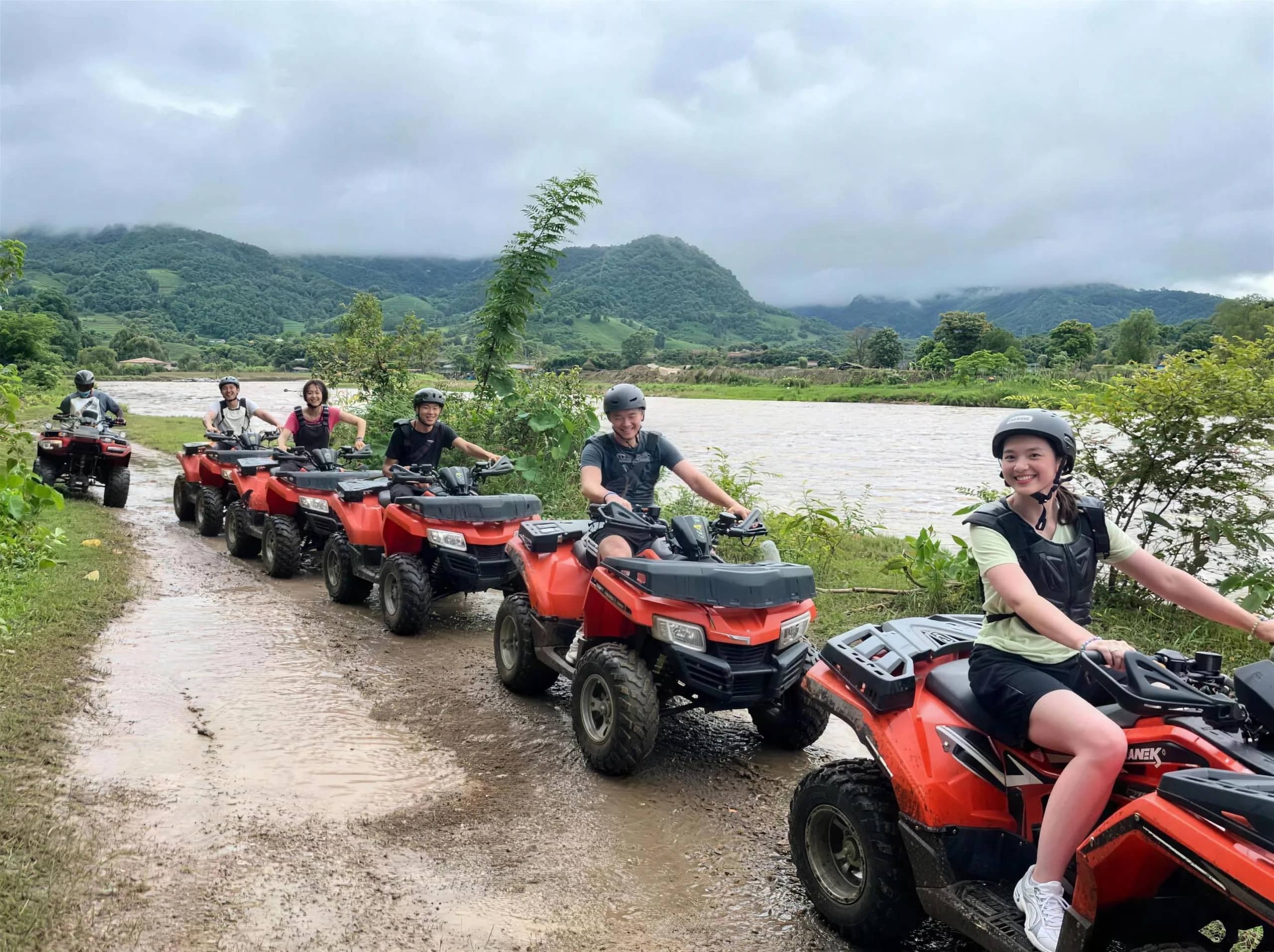 Riders on red ATVs line up along a muddy track beside a river with green mountains behind.