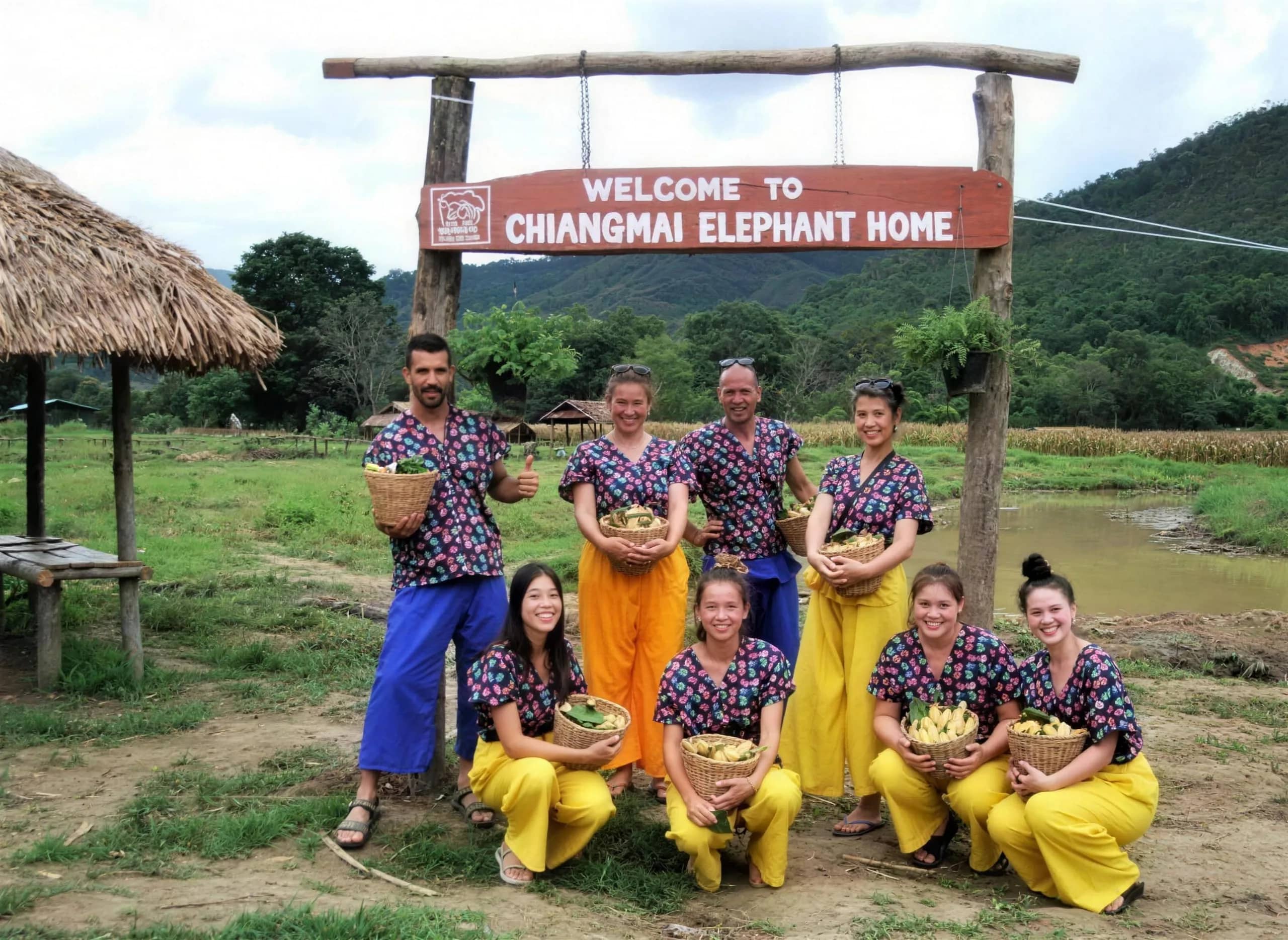 Visitors in patterned tops and baggy trousers pose with baskets under the Chiangmai Elephant Home sign