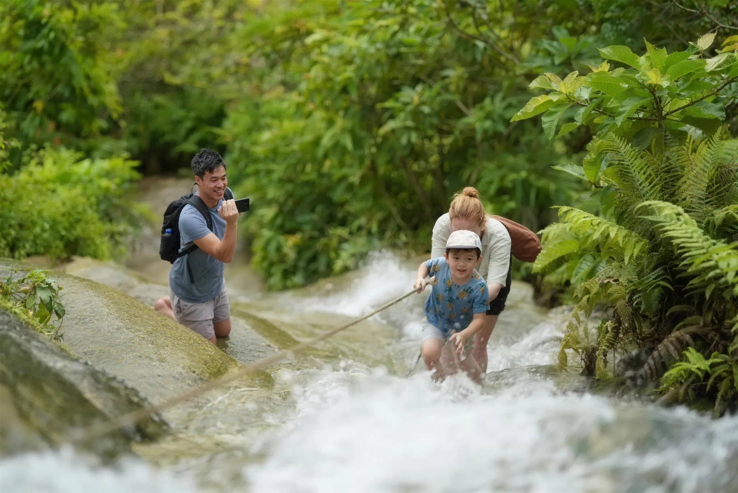 A parent and child hold a rope while climbing the sticky waterfall as another visitor takes photos.