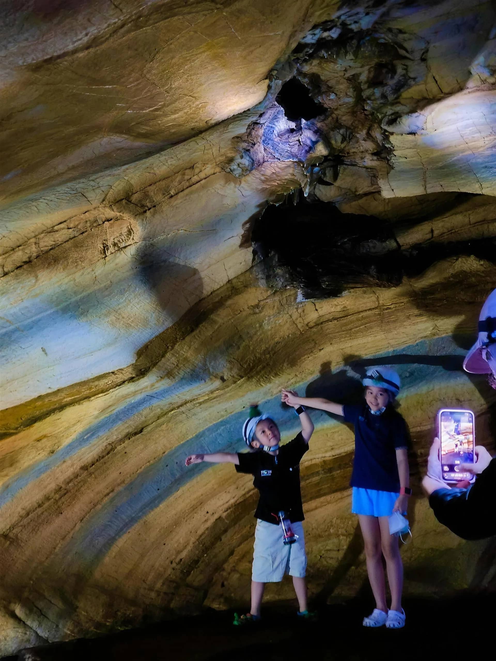 Two children in helmets point at the striped and swirling rock walls inside a cave