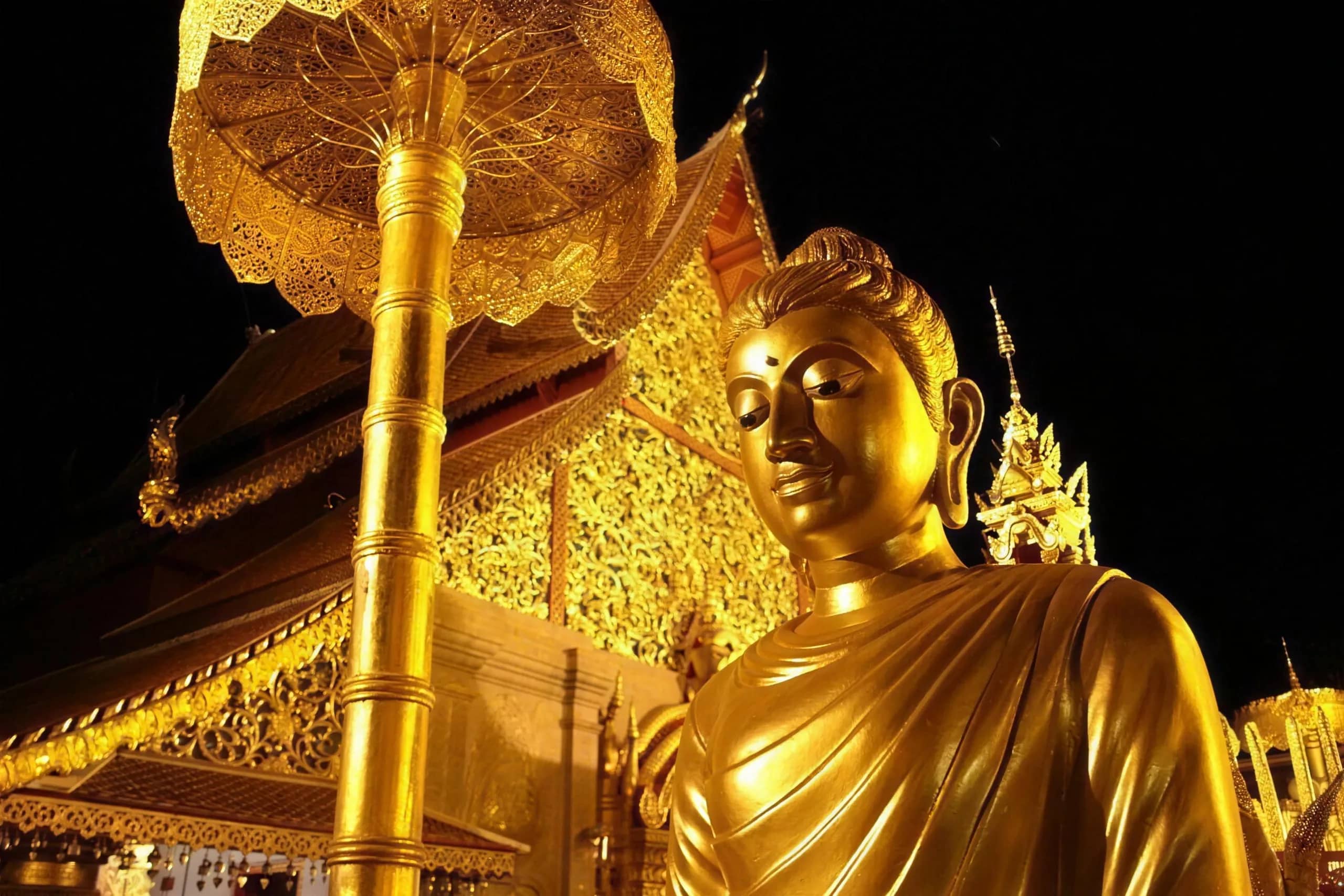 A golden Buddha statue stands under a golden parasol beside an ornate temple hall at night