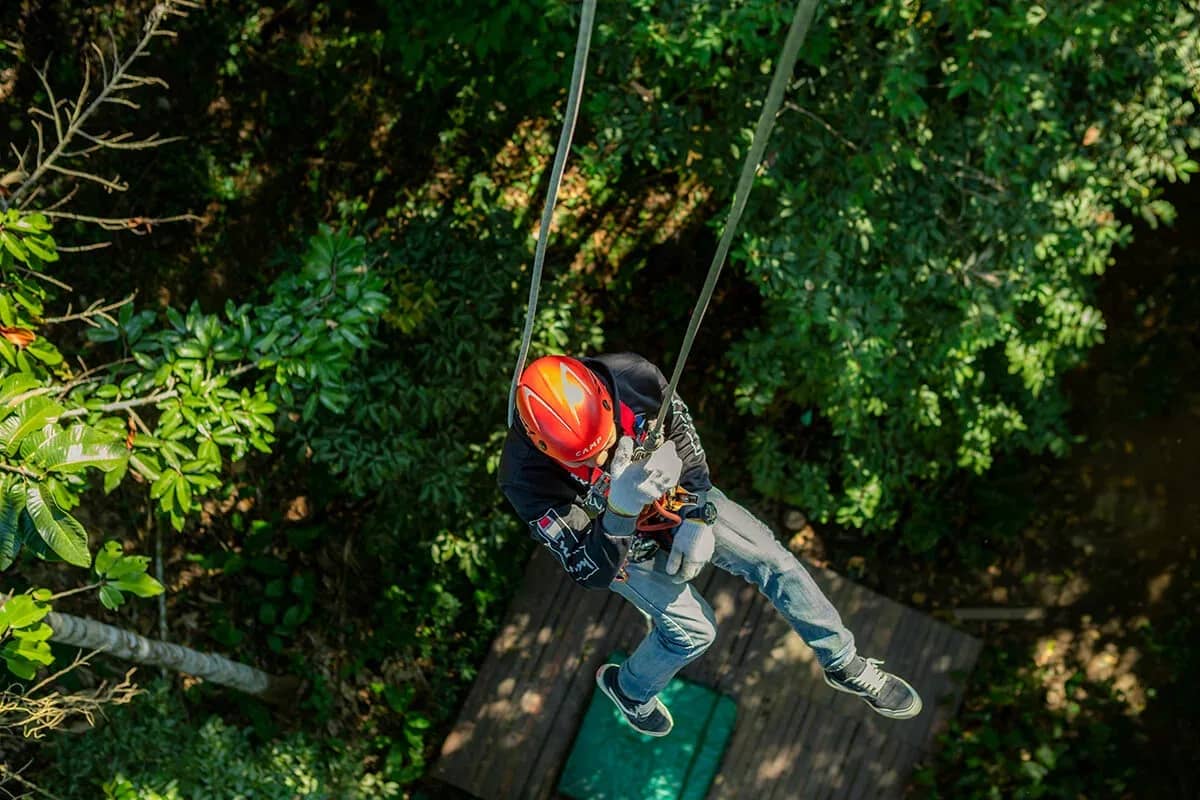A rider in an orange helmet steps off a wooden treetop platform onto the zipline.